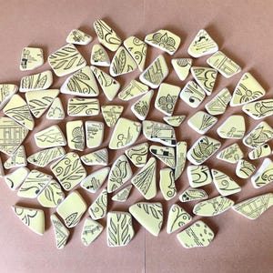 May include: A collection of irregularly shaped, off-white ceramic pieces with black line drawings. The designs include floral, geometric, and abstract patterns. The pieces are scattered on a light brown surface.