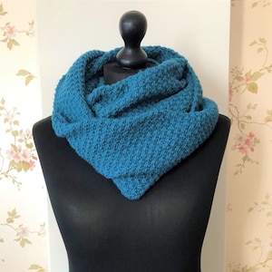 May include: A deep teal infinity scarf with a textured, knitted pattern draped around a black mannequin. The scarf features a consistent, dense stitch throughout, creating a voluminous and layered appearance around the neck area of the display form.