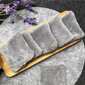 Puede incluir: Seis almohadillas de desmaquillaje reutilizables grises en una bandeja de madera, con una almohadilla adicional en una superficie gris.