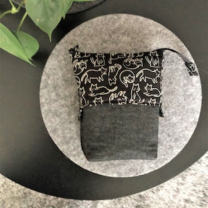 May include: A black zippered pouch with a white cat print design. The pouch is sitting on a gray circle.