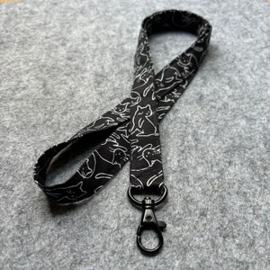 Peut inclure: Un cordon en tissu noir orné d'un motif répétitif de chats en lignes blanches dans diverses positions. Le cordon est constitué d'une boucle continue de tissu et comprend un mousqueton pivotant en métal noir fixé à l'extrémité inférieure.