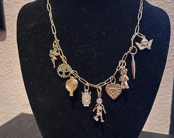 May include: A gold chain necklace with multiple charms, including a hot air balloon, an owl, a heart, a teddy bear, a feather, a fairy, and a tree.