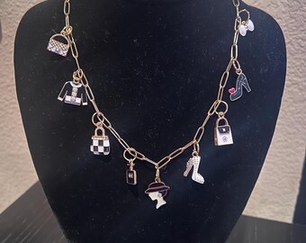 Può includere: Una collana a catena dorata con più ciondoli, tra cui una borsa a scacchi bianco e nero, una borsa a mano bianco e nero, un maglione nero, una scarpa nera con un cuore rosso, una scarpa bianca con strass, un flacone di profumo e una silhouette di una donna con un cappello.