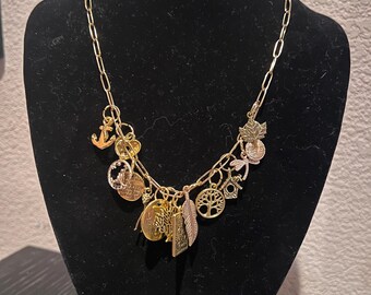 May include: A gold-colored charm necklace displayed on a black mannequin. The necklace features various charms, including an anchor, a tree of life, a feather, and a maple leaf. The charms are in gold and bronze tones, creating a layered effect.