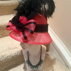 May include: A pink and black hat with a wide brim, adorned with black feathers, bows, and roses. The hat is displayed on a mannequin head, paired with a turquoise and silver necklace. The hat is a stylish accessory.