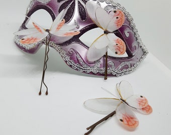 May include: Three white silk butterfly hair clips with pink accents. The butterflies are attached to bobby pins. The butterflies are arranged on a purple and silver masquerade mask.