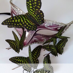 May include: A set of six green and black silk butterfly hair clips. The butterflies have a spotted pattern and are arranged around a purple and silver masquerade mask.