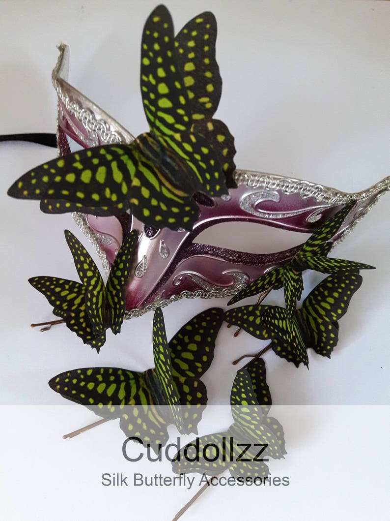 May include: A set of six green and black silk butterfly hair clips. The butterflies have a spotted pattern and are arranged around a purple and silver masquerade mask.