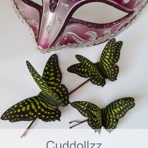 May include: Three green and black silk butterfly hair clips with gold wire stems. The butterflies have a spotted pattern. The image also includes a pink and silver masquerade mask.