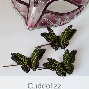 May include: Three green and black silk butterfly hair clips. The butterflies have a spotted pattern and are arranged on a white surface. The text "Cudddollzz Silk Butterfly Accessories" is at the bottom of the image.