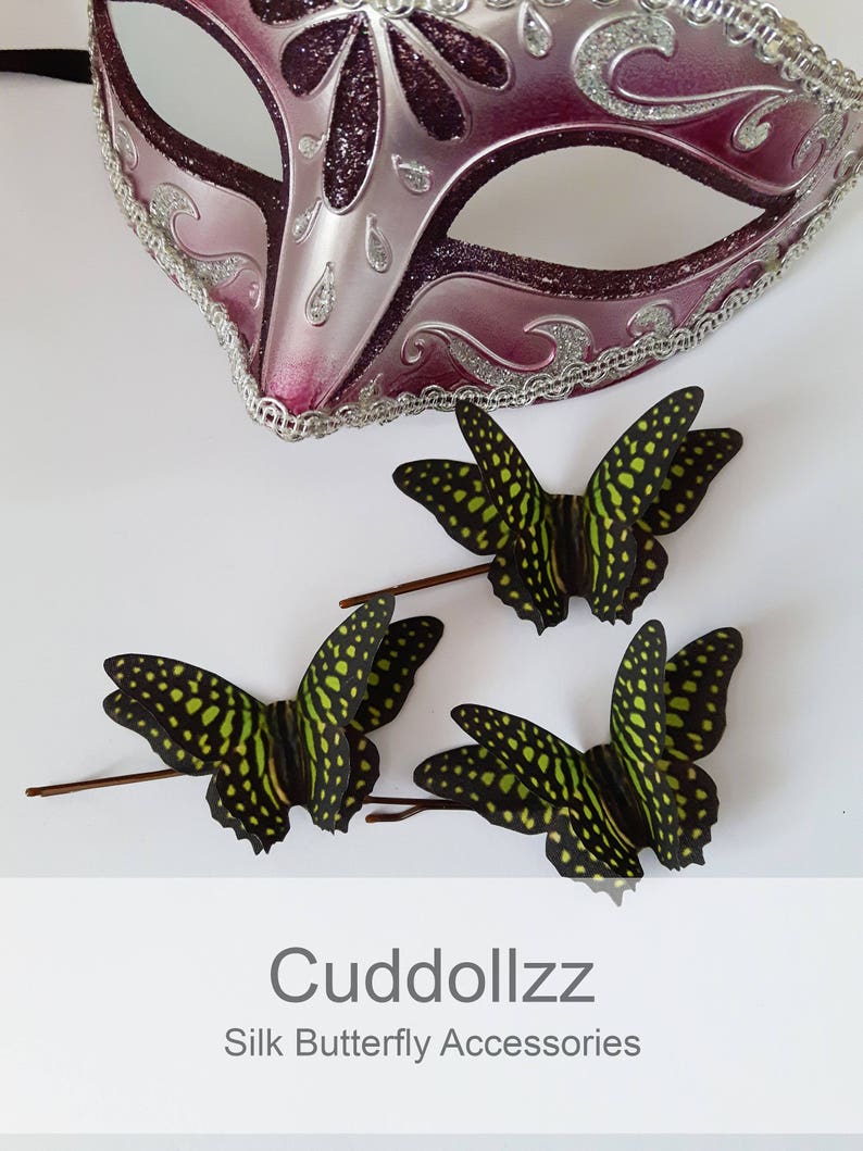 May include: Three green and black silk butterfly hair clips. The butterflies have a spotted pattern and are arranged on a white surface. The text "Cudddollzz Silk Butterfly Accessories" is at the bottom of the image.