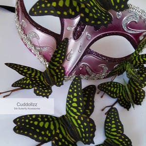 May include: A pink masquerade mask with silver glitter and a decorative border.  The mask is adorned with six black and green silk butterfly hair clips.  The text "Cuddollzzz Silk Butterfly Accessories" is visible below the mask.
