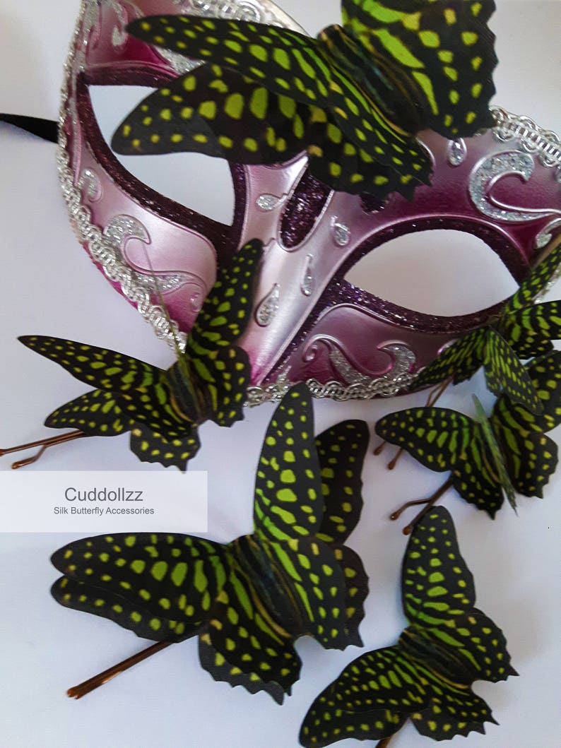 May include: A pink masquerade mask with silver glitter and a decorative border.  The mask is adorned with six black and green silk butterfly hair clips.  The text "Cuddollzzz Silk Butterfly Accessories" is visible below the mask.