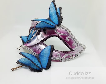 May include: A purple and silver masquerade mask with blue silk butterfly hair clips. The mask has a silver glitter finish and intricate details. The butterflies are attached to bobby pins.