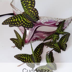 May include: A set of six green and black silk butterfly hair clips. The butterflies are arranged around a pink and silver masquerade mask. The text "Cuddollzz Silk Butterfly Accessories" is at the bottom of the image.