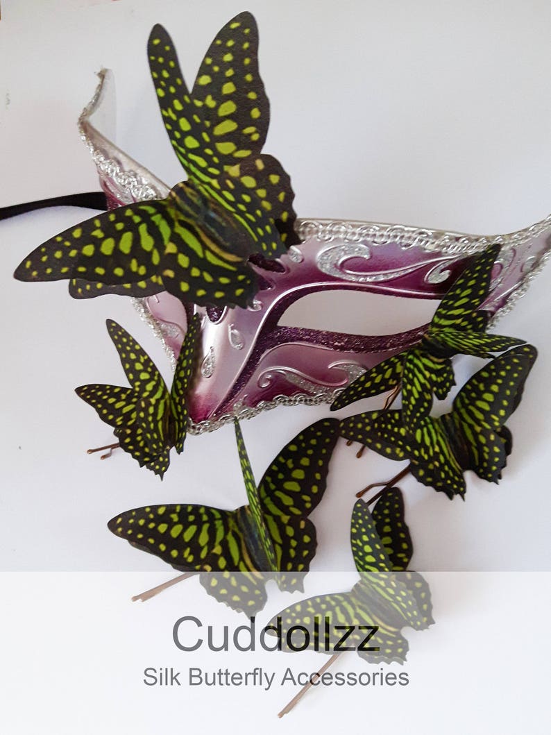 May include: A set of six green and black silk butterfly hair clips. The butterflies are arranged around a pink and silver masquerade mask. The text "Cuddollzz Silk Butterfly Accessories" is at the bottom of the image.