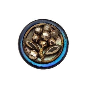 May include: A circular button featuring a dark blue iridescent rim and a central floral motif. The flower is composed of faceted metallic beads and two smooth, dark, leaf-shaped accents set against a dark, reflective background.