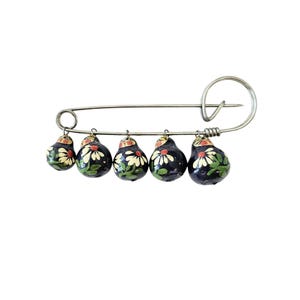 May include: A silver safety pin with five dangling wooden ornaments. The ornaments are painted black with white flowers and green leaves.