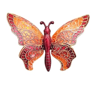 Könnte beinhalten: Eine rote und orangefarbene Schmetterlingsbrosche mit filigranen goldenen Details. Der Schmetterling hat ein detailliertes Design auf seinen Flügeln und einen roten Körper.