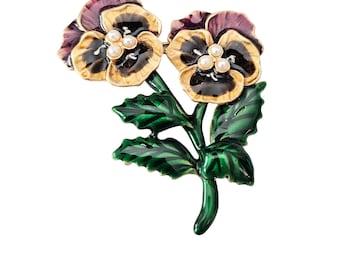May include: A floral brooch featuring two pansy flowers with purple, gold, and black petals. The brooch has faux pearl accents and green leaves and stem. The brooch is made of metal and enamel.