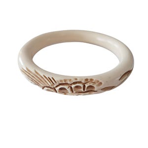 May include: Cream-coloured bangle bracelet featuring carved floral and geometric patterns. The bracelet is a solid, circular band with a smooth, rounded exterior and detailed carvings in a contrasting brown hue.
