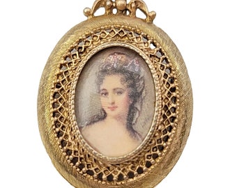 Könnte beinhalten: Goldenes ovales Medaillon mit einem filigranen Design. Das Medaillon zeigt ein Miniaturporträt einer Frau mit einem weißen Spitzenkragen und einem Blumenhaarschmuck.