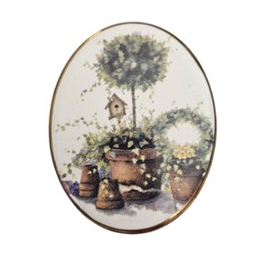 Könnte beinhalten: Ovale dekorative Plakette mit goldfarbenem Rand. Das Bild zeigt Topfpflanzen, darunter einen Formschnittbaum, ein Vogelhaus und einen Kranz. Die Farbpalette umfasst Grün-, Braun- und Weißtöne und schafft eine ruhige Gartenszene.