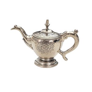 Könnte beinhalten: Eine silberne Teekanne mit aufwendigen Details. Die Teekanne hat einen gebogenen Ausguss, einen dekorativen Griff und einen Deckel mit einem Knauf. Der Körper der Teekanne ist mit einem floralen Design und einem Wappen graviert. Sie steht auf einem runden Sockel.