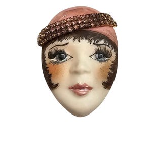 May include: A vintage porcelain face with a pink hat and a sparkling rhinestone band. The face has detailed features, including dark eyelashes, blue eyes, and rosy cheeks. The lips are painted a soft pink, and the overall design is reminiscent of a flapper era.