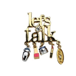 Op de afbeelding: Goudkleurige metalen broche met de woorden "let's talk" en bungelende bedels. Bedels zijn onder meer een parfumflesje, nagellak, een potje, lippenstift en een ovale bedel met het woord "Avon". Een heldere strass-steen is gezet in het woord "talk".