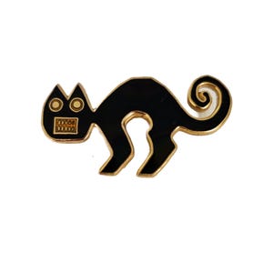 Puede incluir: Pin de esmalte negro de un gato estilizado con cola enroscada. El gato tiene un contorno dorado y una cara dorada con un patrón de cuadrícula.