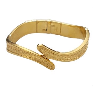 May include: A gold-toned bangle bracelet with a textured, wave-like design. The bracelet has a hinged clasp.