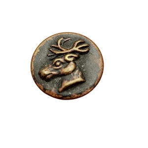 May include: A round button featuring a raised, metallic-toned relief of a deer head with antlers. The button has a dark, weathered finish with copper-colored highlights along the edges and the embossed deer design.