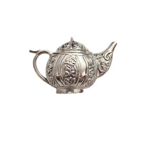 May include: A silver-colored teapot with intricate embossed floral and scrollwork designs. The teapot features a curved spout, a rounded body, and a small handle. The lid is also decorated with similar patterns and a small knob.