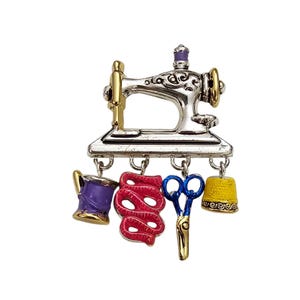 May include: A silver sewing machine brooch with gold accents and purple, red, blue, and yellow hanging charms. The charms include a spool of thread, measuring tape, scissors, and a thimble.
