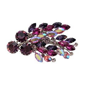 Puede incluir: Broche vintage con diseño floral sobre una base de metal plateado. Presenta cristales morados, rosas e iridiscentes de diversas formas y tamaños. Los cristales están dispuestos para asemejarse a hojas y flores.