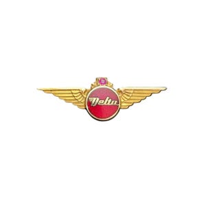 Puede incluir: Un pin dorado de Delta Airlines con alas. El pin presenta un círculo rojo con la palabra "Delta" en escritura blanca. Una pequeña gema rosa se encuentra encima del círculo.