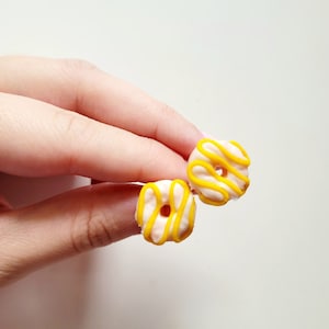 Könnte beinhalten: Ein Paar Donut-Ohrringe mit weißem Zuckerguss und gelber Glasur.
