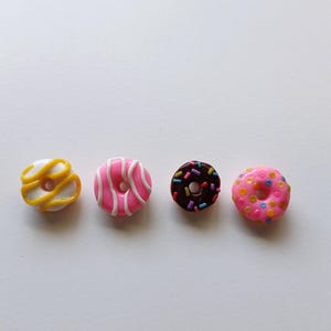 May include: Four miniature donuts with different toppings. A white donut with yellow icing, a pink donut with white stripes, a brown donut with colourful sprinkles, and a pink donut with yellow and orange sprinkles.