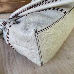 May include: Cream-colored leather handbag with brown stitching and handles. The bag has a textured surface and a rectangular shape. The stitching detail is visible along the edges and handles, adding a decorative element to the design.