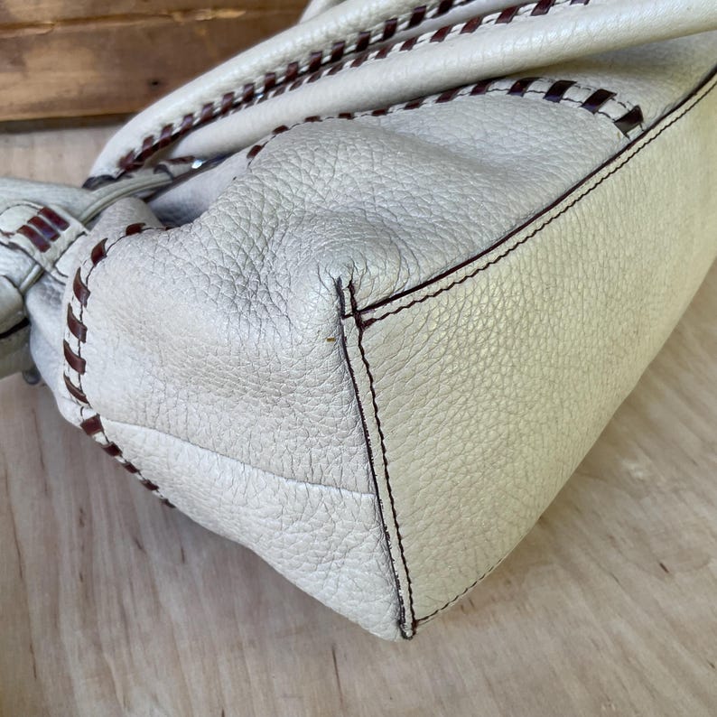 May include: Cream-colored leather handbag with brown stitching and handles. The bag has a textured surface and a rectangular shape. The stitching detail is visible along the edges and handles, adding a decorative element to the design.