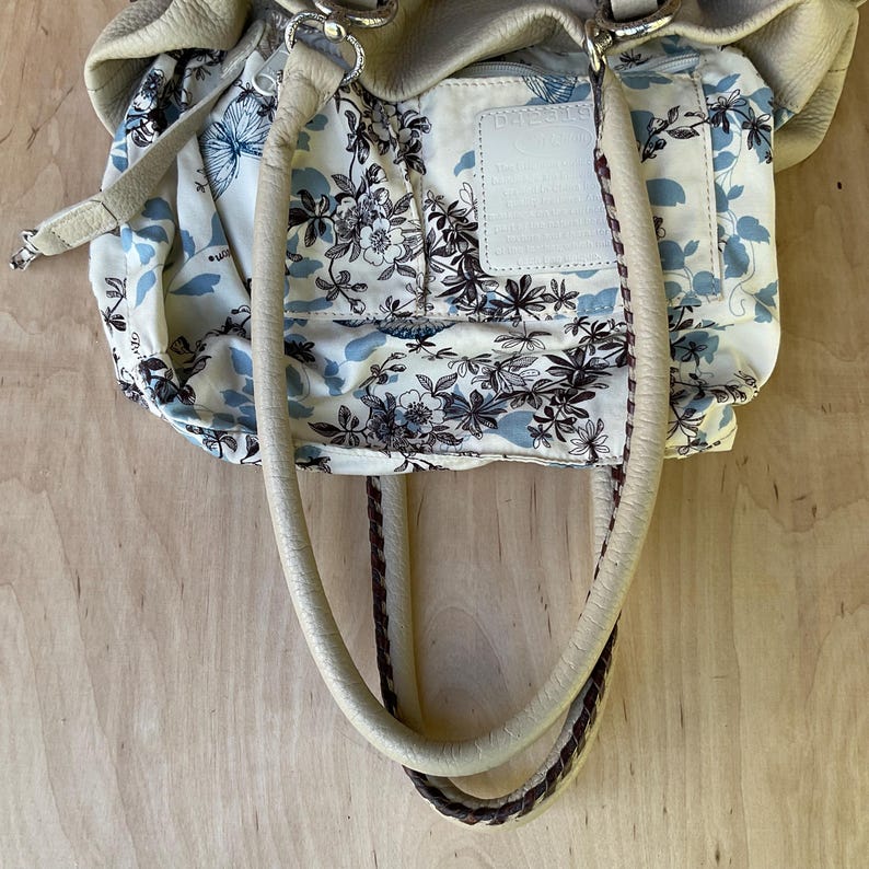 May include: Cream-colored leather handbag with a floral patterned interior. The bag features a light blue and black floral design on a cream background. The handles are cream-colored with a braided brown accent. Silver-toned hardware completes the design.