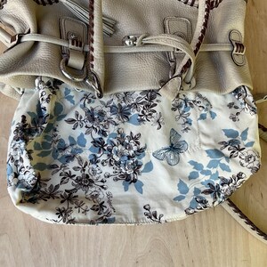 May include: Cream-colored leather handbag with a floral and butterfly patterned lining. The bag features braided accents, silver-tone hardware, and a shoulder strap. The interior fabric has a blue and white floral design.