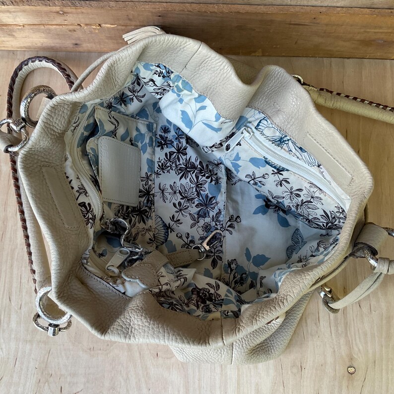 May include: Cream-colored leather handbag with a floral patterned interior. The bag features a top handle, a shoulder strap, and multiple compartments. The interior lining has a blue and white floral design with butterflies.