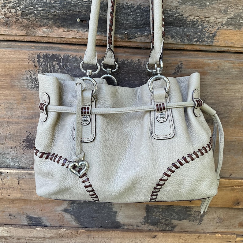 May include: A light beige leather handbag with brown stitching and braided handles. The bag features silver-tone hardware, a heart-shaped charm, and a drawstring closure. The stitching detail adds a decorative element to the bag's design.