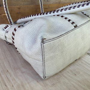 May include: Cream-colored leather handbag with dark brown stitching and accents. The bag features a textured surface and a stitched handle. The stitching detail is visible on the handle and around the edges of the bag.