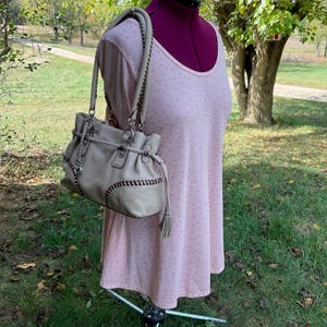 May include: A light gray leather handbag with stitched detailing and silver-tone hardware. The bag has two handles with braided accents and a tassel. The bag is displayed on a pink dress form in an outdoor setting.