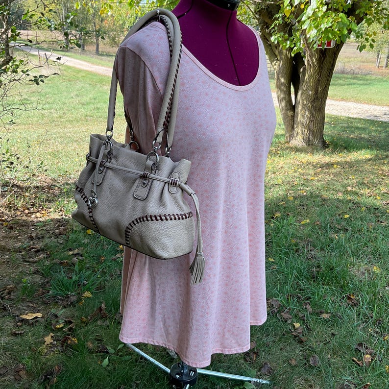 May include: A light gray leather handbag with stitched detailing and silver-tone hardware. The bag has two handles with braided accents and a tassel. The bag is displayed on a pink dress form in an outdoor setting.