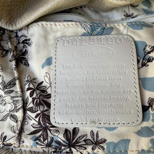May include: Close-up of a Brighton handbag interior. The lining features a floral pattern in black and blue on a cream background. A white leather tag displays the Brighton logo and text about the handbags being crafted from quality leather.