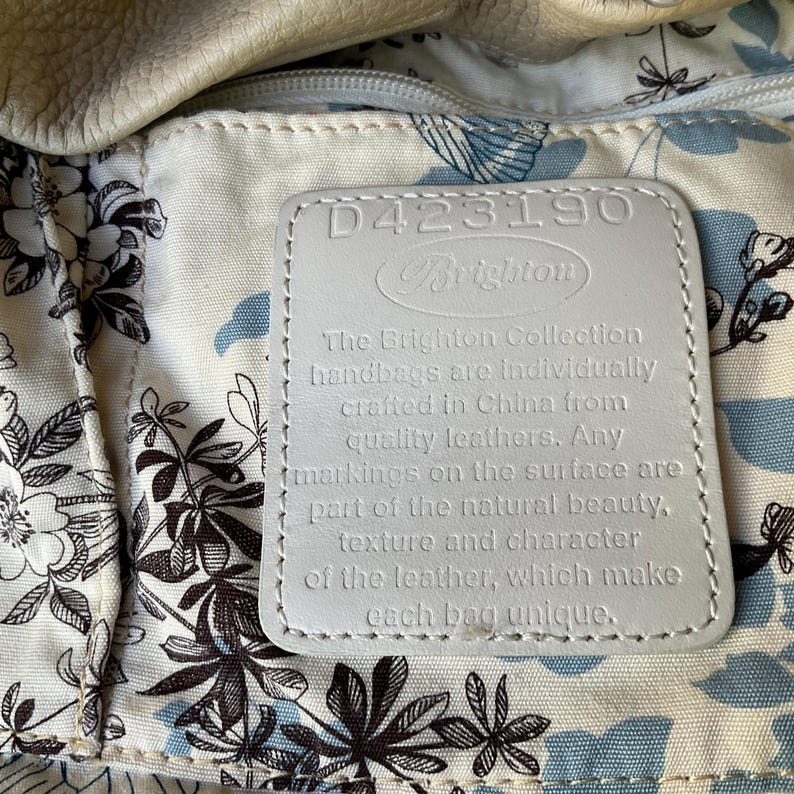 May include: Close-up of a Brighton handbag interior. The lining features a floral pattern in black and blue on a cream background. A white leather tag displays the Brighton logo and text about the handbags being crafted from quality leather.